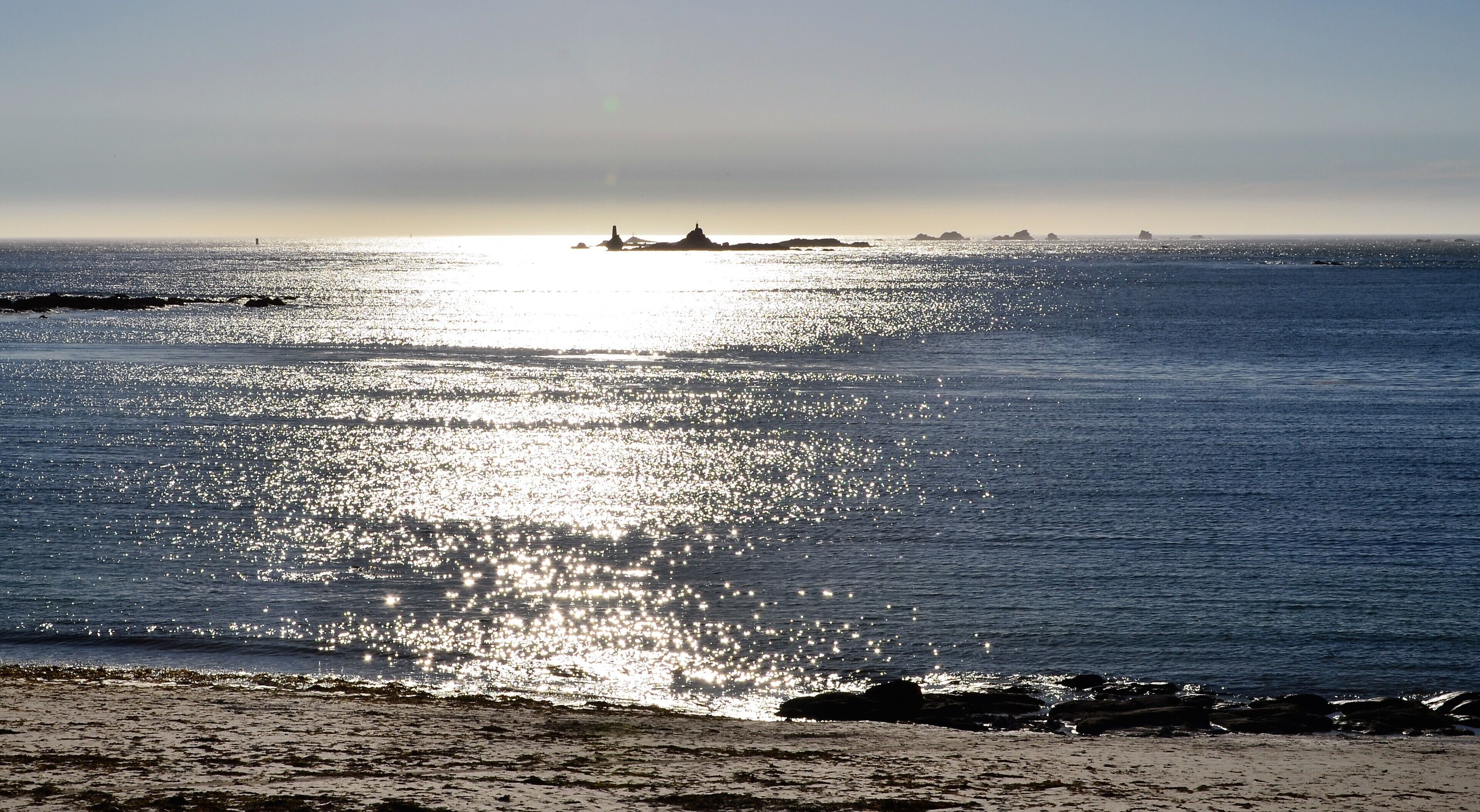 A beach with a calm sea reflecting sunlight, distant islands, and a clear sky.