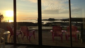 Um terraço com cadeiras e uma mesa, com vista para um corpo de água e terra distante ao pôr do sol.
