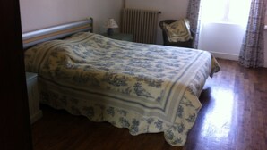Une chambre à coucher comprenant un lit, une table de chevet, une lampe, une chaise, un radiateur et une fenêtre avec des rideaux.