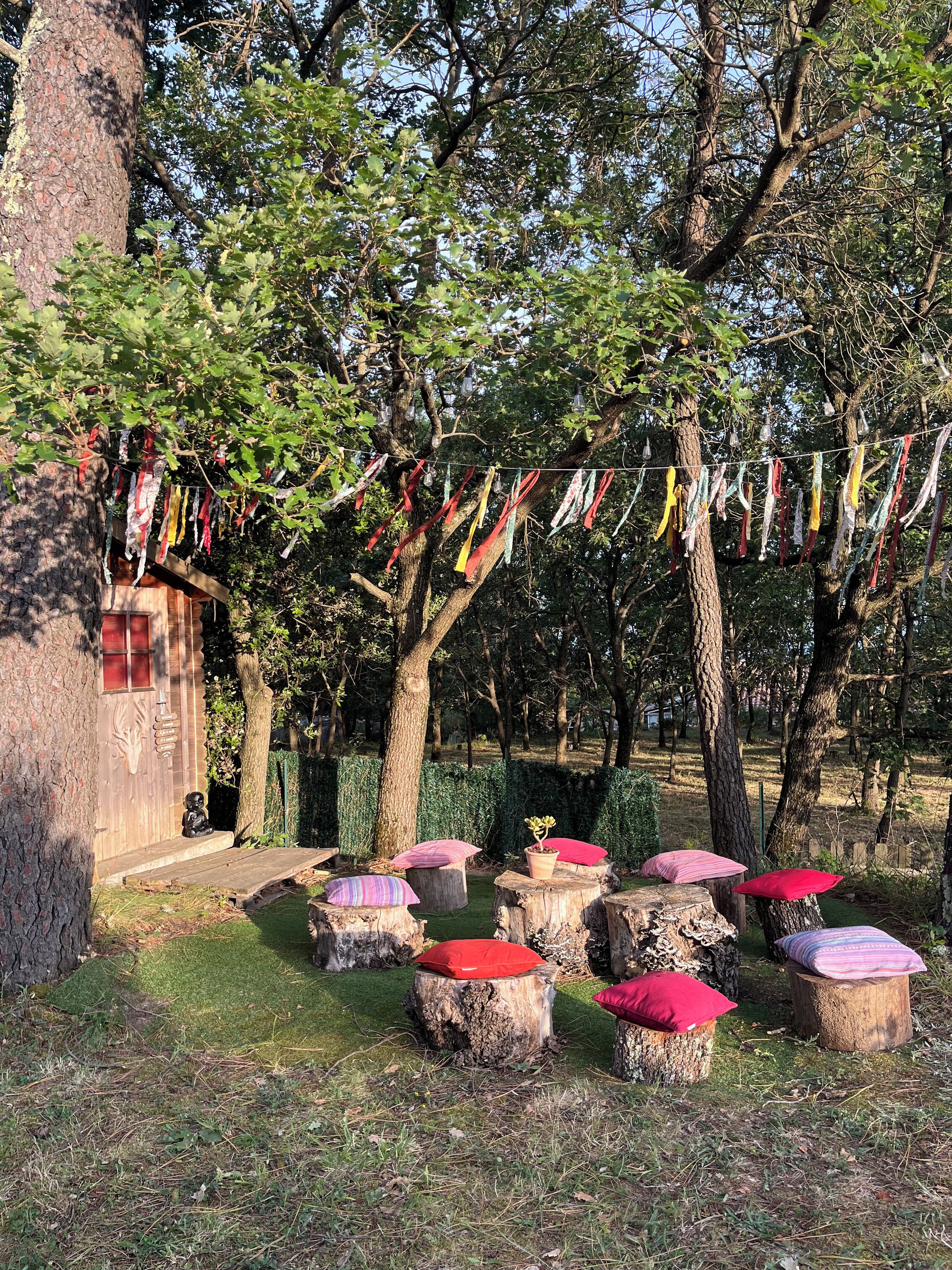 A rustic outdoor setting with a wooden cabin, a clothesline with colorful cloths, and several stools with cushions.