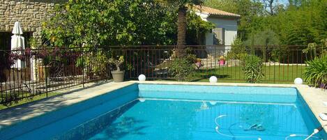 A swimming pool with a tiled edge, surrounded by a metal fence, and adjacent to a stone building with a tiled roof.