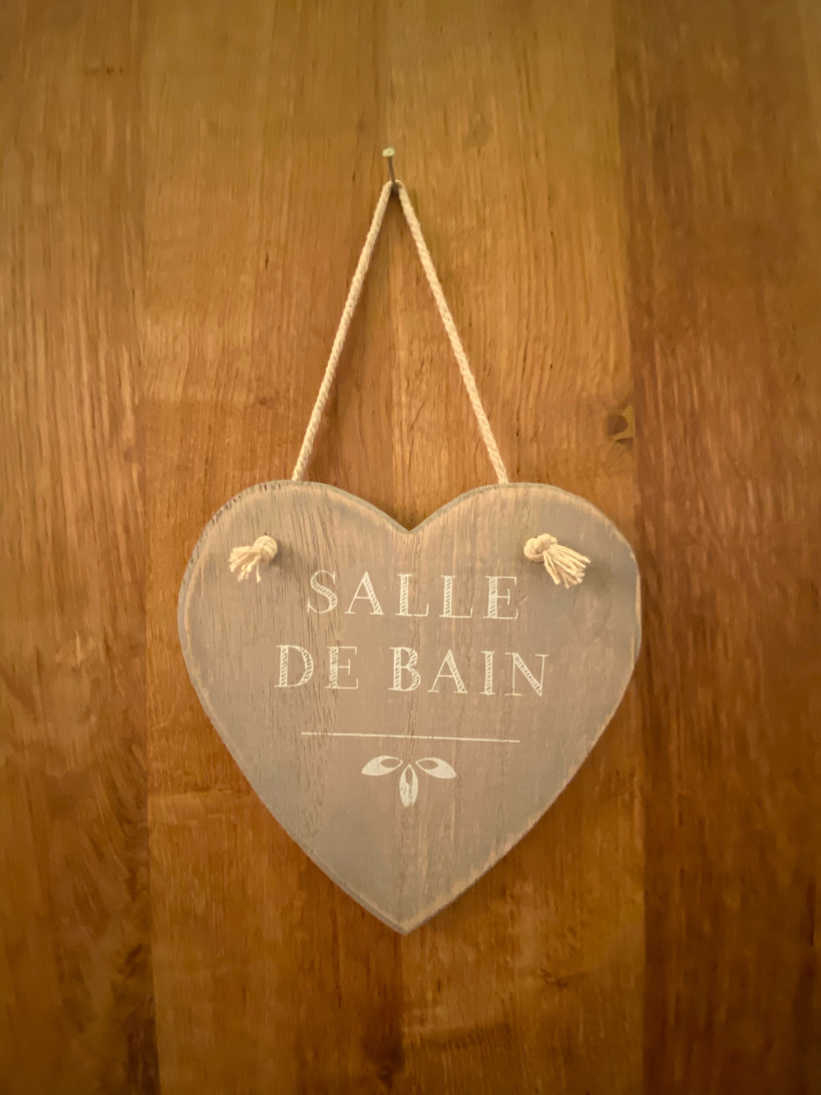 Une enseigne en bois en forme de cœur, portant l’inscription « SALLE DE BAIN », est accrochée à un mur en bois.