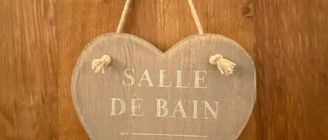 Une enseigne en bois en forme de cœur, portant l’inscription « SALLE DE BAIN », est accrochée à un mur en bois.