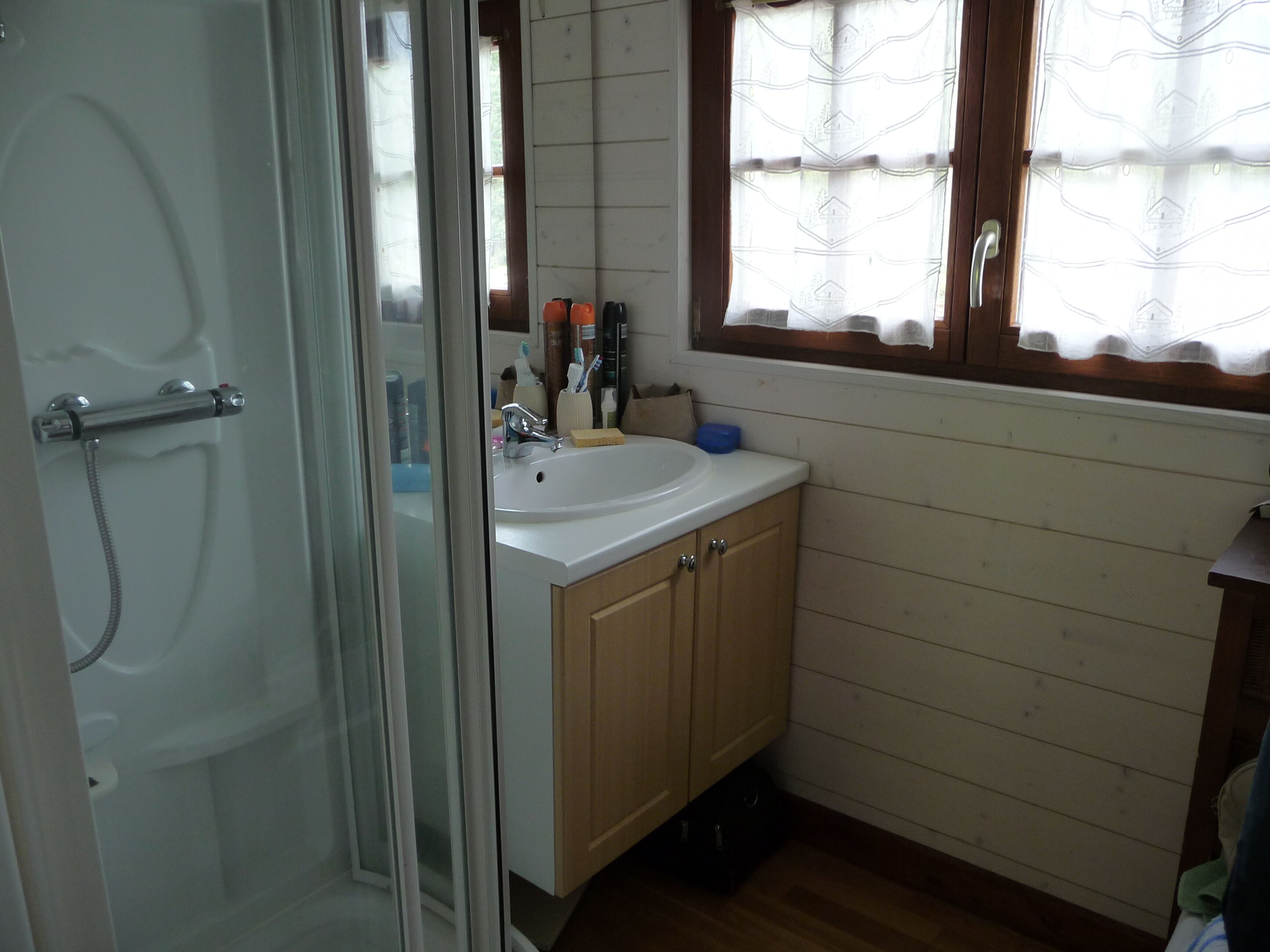 Ein Badezimmer mit Glasduschtür, Waschbecken mit Spiegel darüber, Holzschränken und einem Fenster mit gemustertem Vorhang.