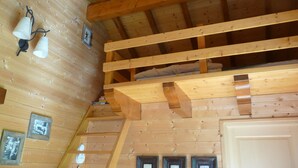 Interior de una cabaña de madera con cama en el altillo, cuadros en las paredes y escalera de madera.