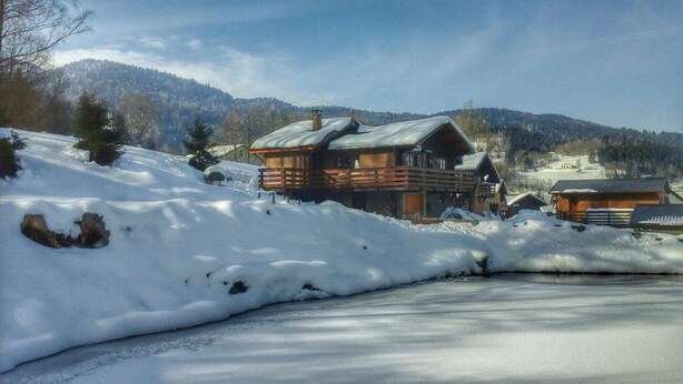 Una cabaña de madera con techo nevado y una terraza, rodeada de un paisaje invernal y montañas.