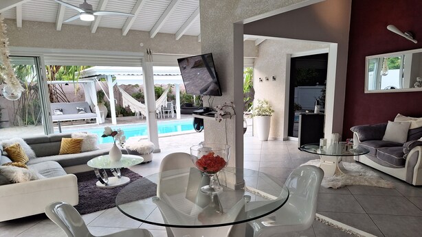 Un salon moderne avec une table en verre, des chaises blanches, un canapé gris et un espace piscine visible à travers les portes vitrées.