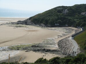 Paisagem costeira com uma praia de areia, um caminho de pedra e uma colina com edifícios.