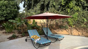 Deux fauteuils de jardin avec des coussins à rayures et un parasol rouge, placés près d’une piscine entourée d’une végétation luxuriante.