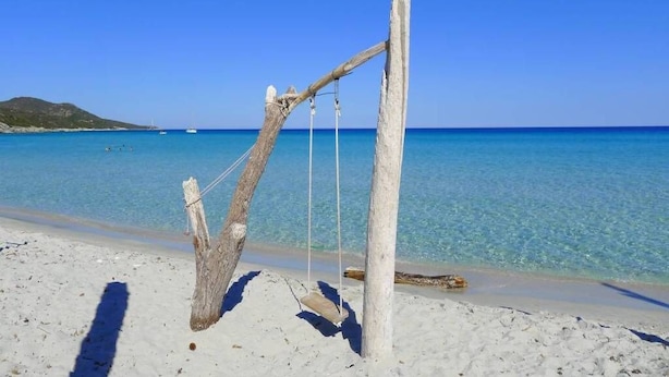 Une plage avec une balançoire faite de poteaux en bois et de cordes, une eau d’un bleu cristallin et une colline qui se dessine à l’horizon.