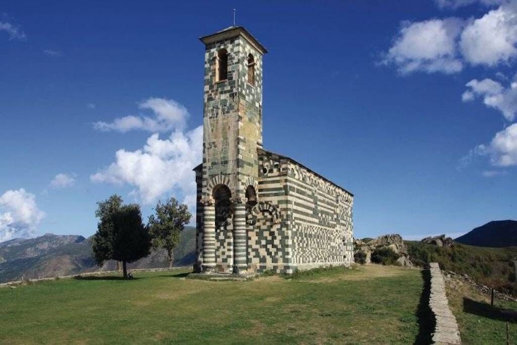 Ein historisches Gebäude mit einem hohen Turm und einer Schachbrettfassade, umgeben von einer Wiese und Bergen im Hintergrund.