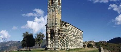 Ein historisches Gebäude mit einem hohen Turm und einer Schachbrettfassade, umgeben von einer Wiese und Bergen im Hintergrund.