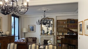 Un vaste salon agrémenté d’un magnifique piano à queue, d’une cheminée, d’un lustre et d’une grande bibliothèque remplie de livres.