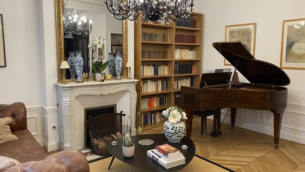 Un salon agrémenté d’un magnifique piano à queue, d’une cheminée, d’un grand miroir et d’une bibliothèque remplie de livres.