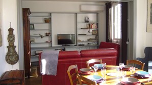 A dining area with a red sofa, wooden dining table, and chairs. There is a television on a shelf and a window with curtains. - House 10 mn walk from the city center with shaded garden, olive trees.  (Uzès)