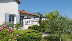 A two-story house with a covered porch, a garden with various plants and flowers, and a clear blue sky. - House 10 mn walk from the city center with shaded garden, olive trees.  (Uzès)
