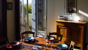 A dining area with a table set for four, a window with a view of greenery, and a cabinet with a basket on top. - House 10 mn walk from the city center with shaded garden, olive trees.  (Uzès)