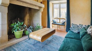 Una sala de estar con chimenea de piedra, plantas verdes, un banco de madera, un sofá verde y un comedor con una silla y una mesa.