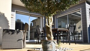 Un patio con un árbol en maceta, mobiliario exterior y un ventanal corredero que da a una zona cubierta.