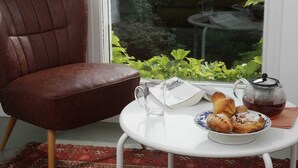 Ein brauner Ledersessel, ein weißer Tisch mit Teekanne, einem Tablett Gebäck und ein Fenster mit Grünpflanzen im Hintergrund.