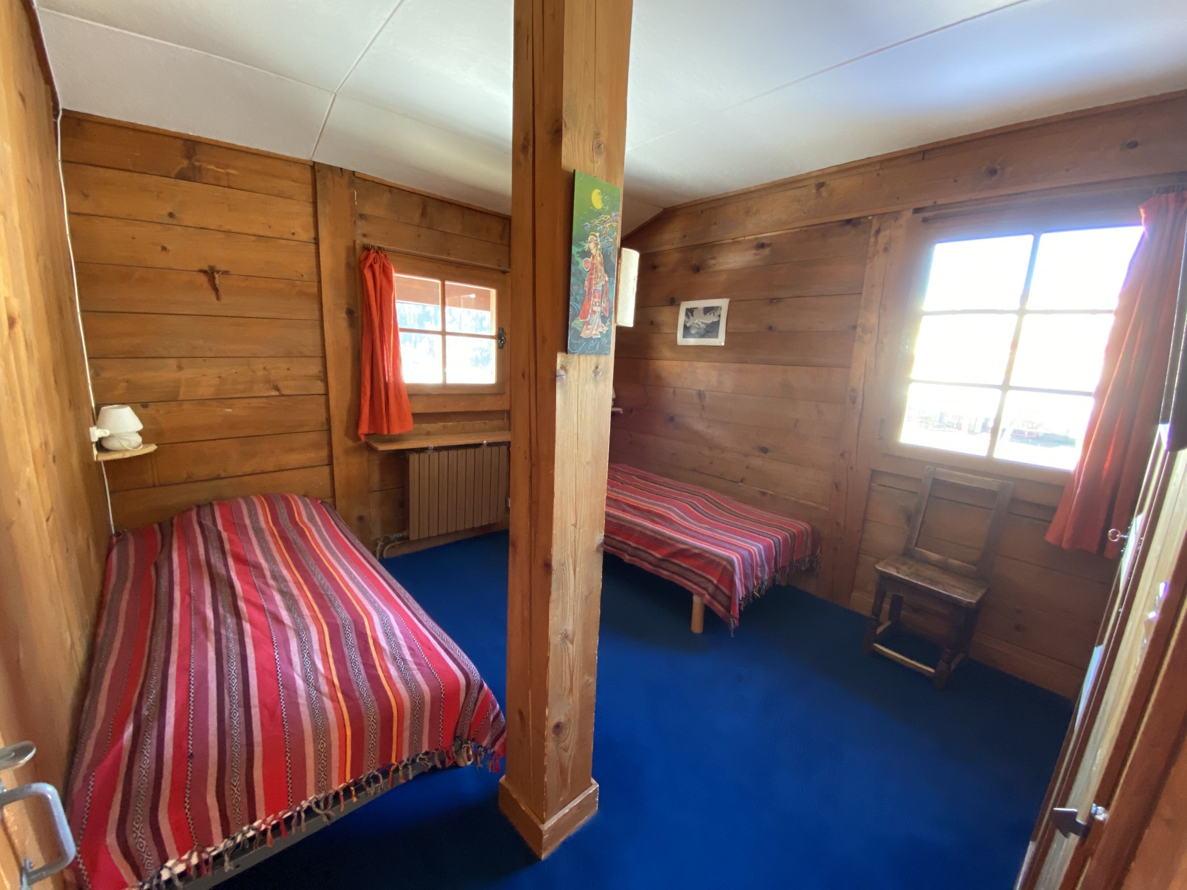 Una habitación tipo cabaña con dos camas, paredes de madera, alfombra azul, una ventana con cortinas y una mesita.