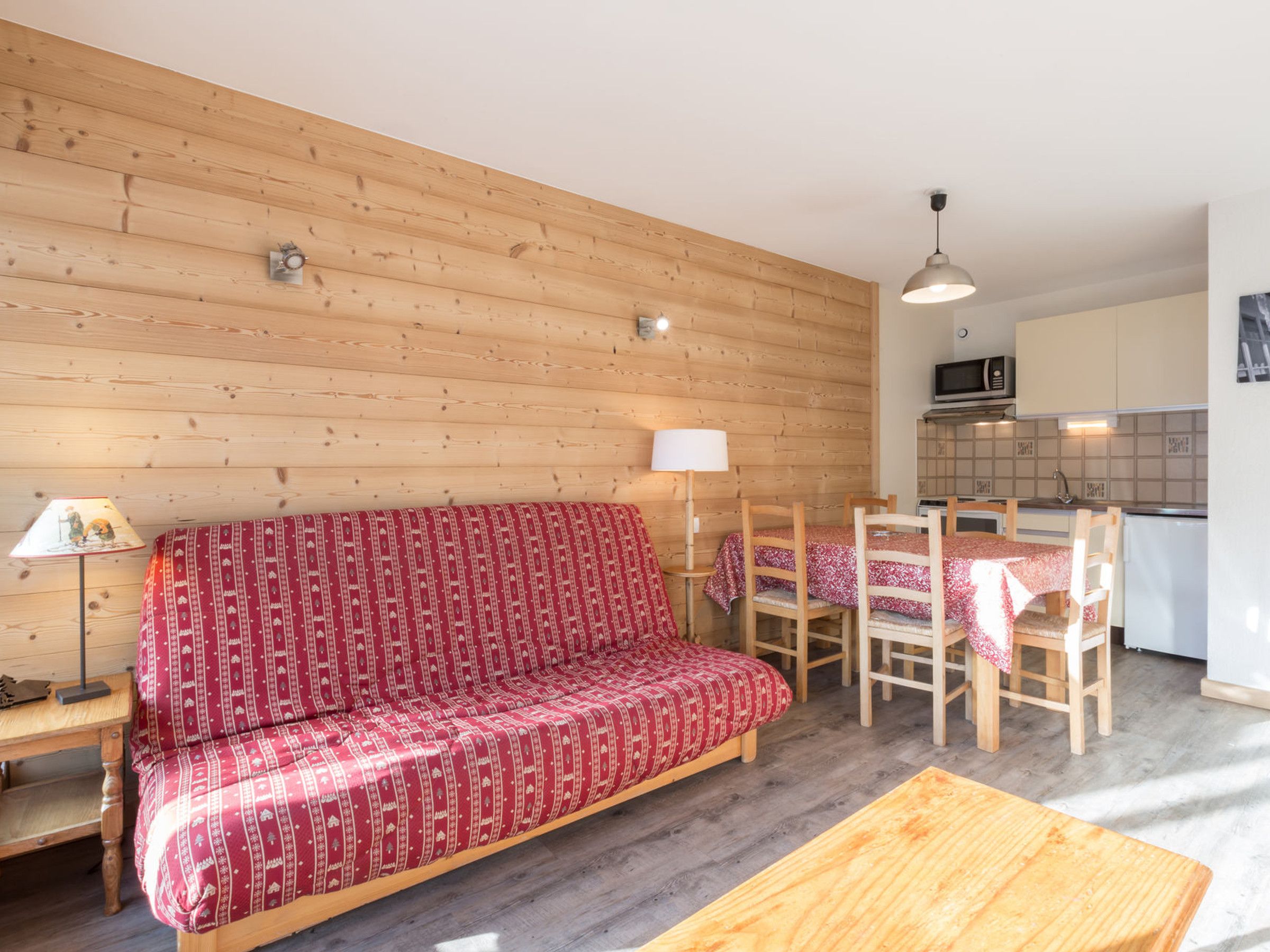 A cozy room with a wooden sofa, a dining table with chairs, a lamp, and a kitchen area with cabinets and a microwave.