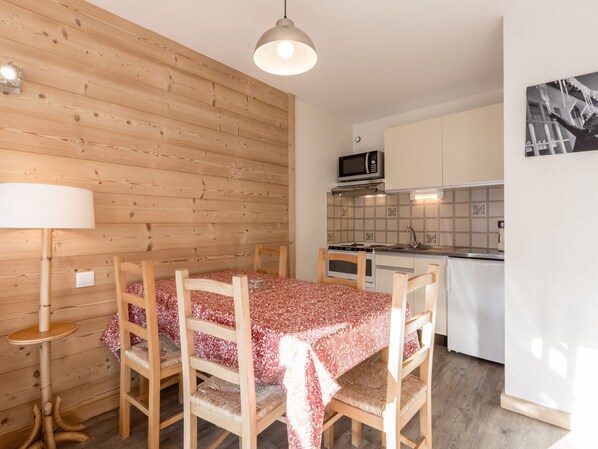 A compact kitchen with a microwave, stove, and sink. There is a dining table with chairs and a lamp. The room has wooden paneling on the walls.