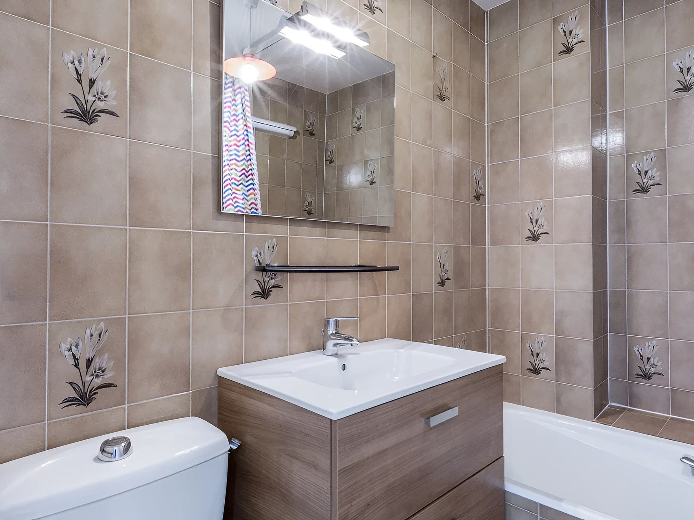 A bathroom with beige tiles, a white sink, a wooden vanity, a mirror, and a shower.