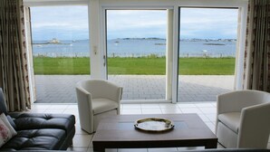 Una sala de estar moderna con un sofá, sillones y una mesa de centro. Amplias puertas de vidrio dan a un patio con vista al mar y una zona ajardinada.