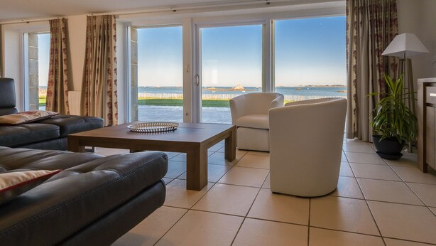 Una sala de estar moderna con un sofá de cuero, un sillón blanco, una mesa de centro de madera y vistas al mar a través de grandes ventanales.