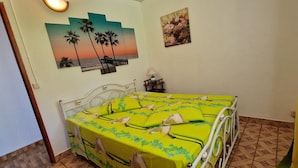 A bedroom with a bed covered in a green floral bedspread, a white metal bed frame, a wall-mounted lamp, and a painting of a sunset with palm trees. - Detached house rated 03 stars, comfortable, equipped, with Wifi (deshaies)