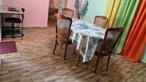 A dining area with a table covered in a patterned tablecloth, surrounded by four wooden chairs with woven seats. A laptop is open on a desk to the left. - Detached house rated 03 stars, comfortable, equipped, with Wifi (deshaies)
