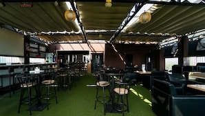 Espaço interior com mesas e cadeiras dispostas para refeições, piso verde e teto alto com vigas aparentes e luminárias pendentes.