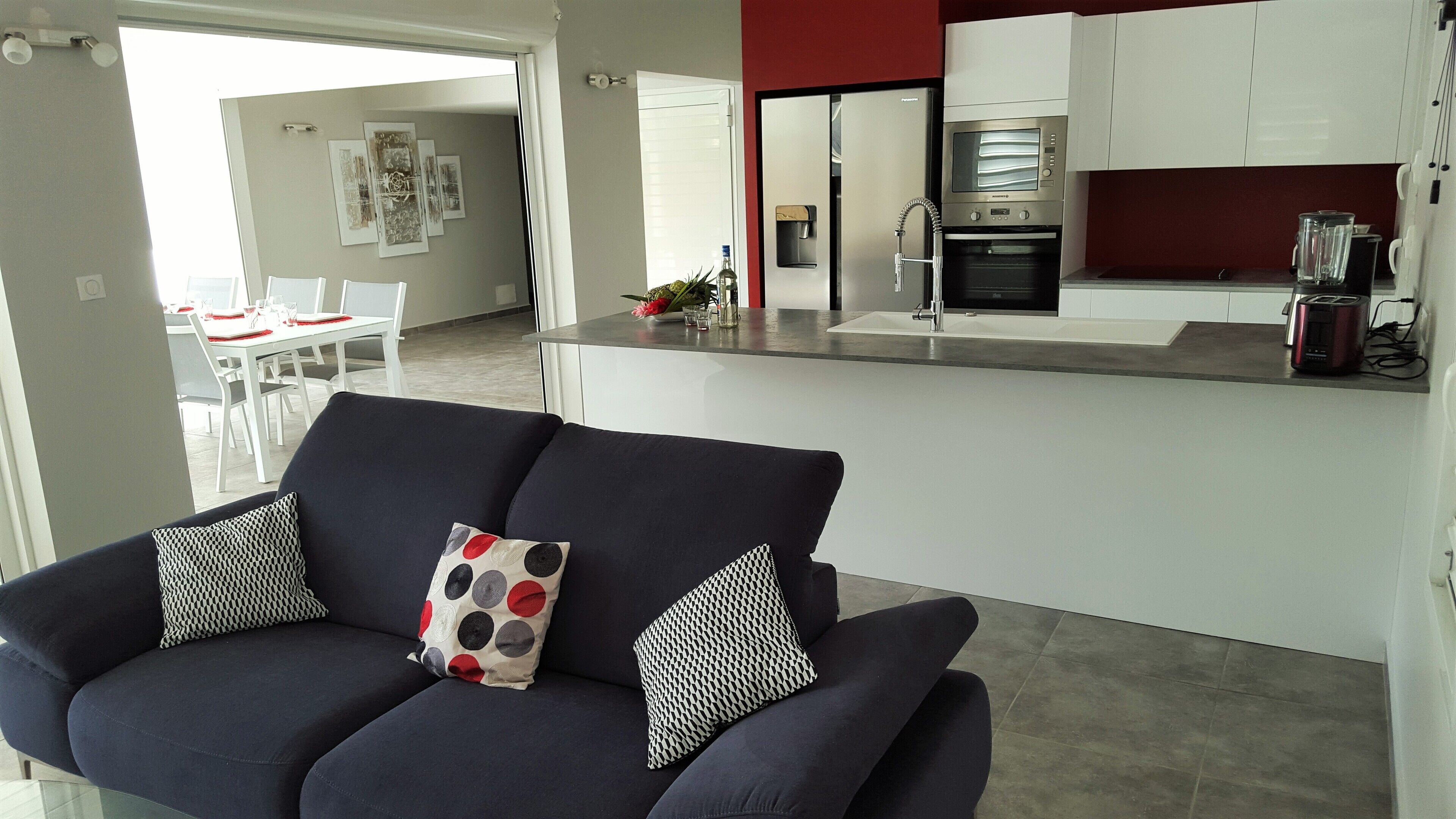 A modern living room with a dark sofa and patterned cushions, a kitchen with white cabinets and a built-in oven, and a dining area with a white table and chairs.