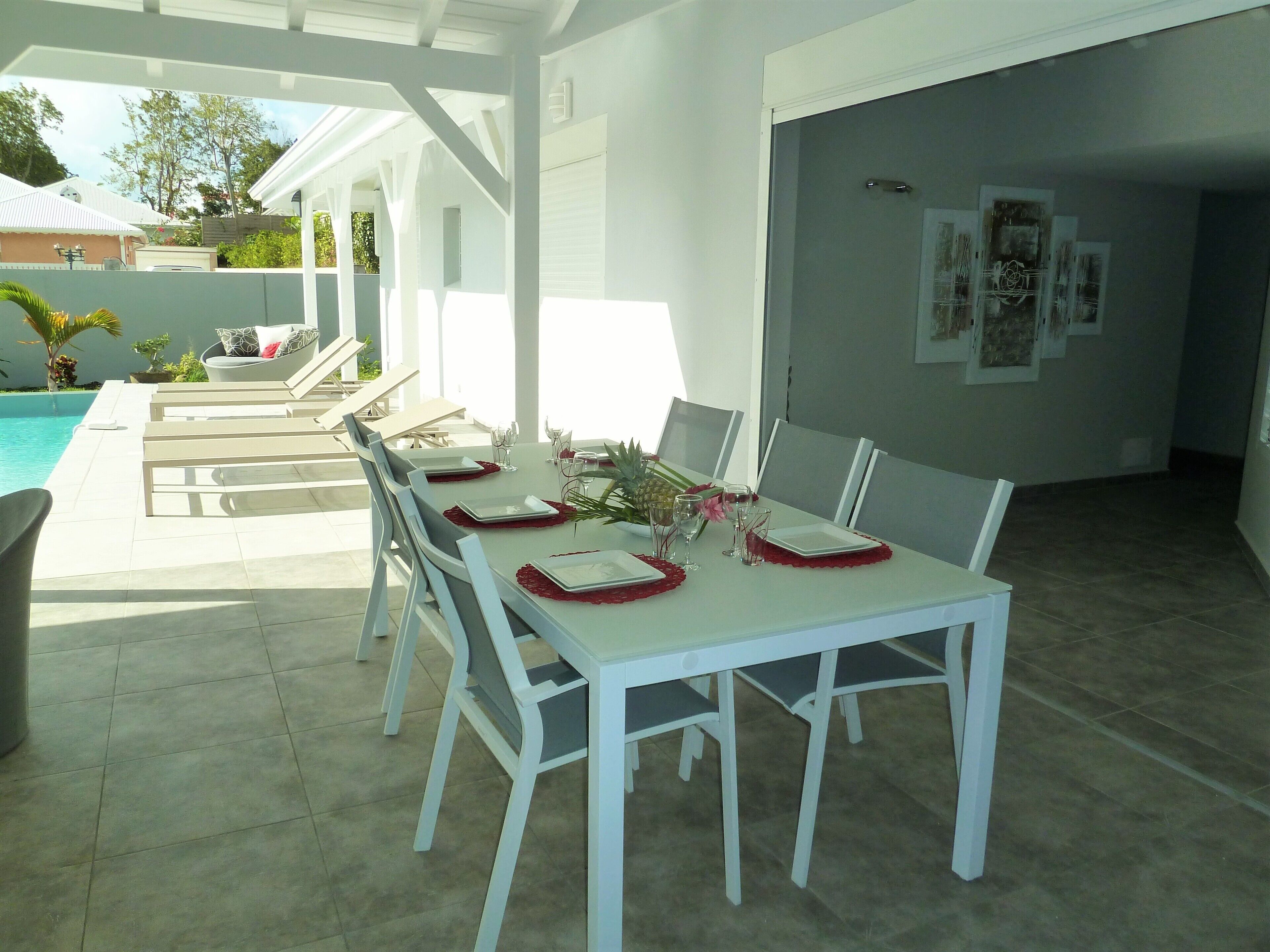 A dining area with white tables and chairs, a pool in the background, and a covered outdoor seating area.
