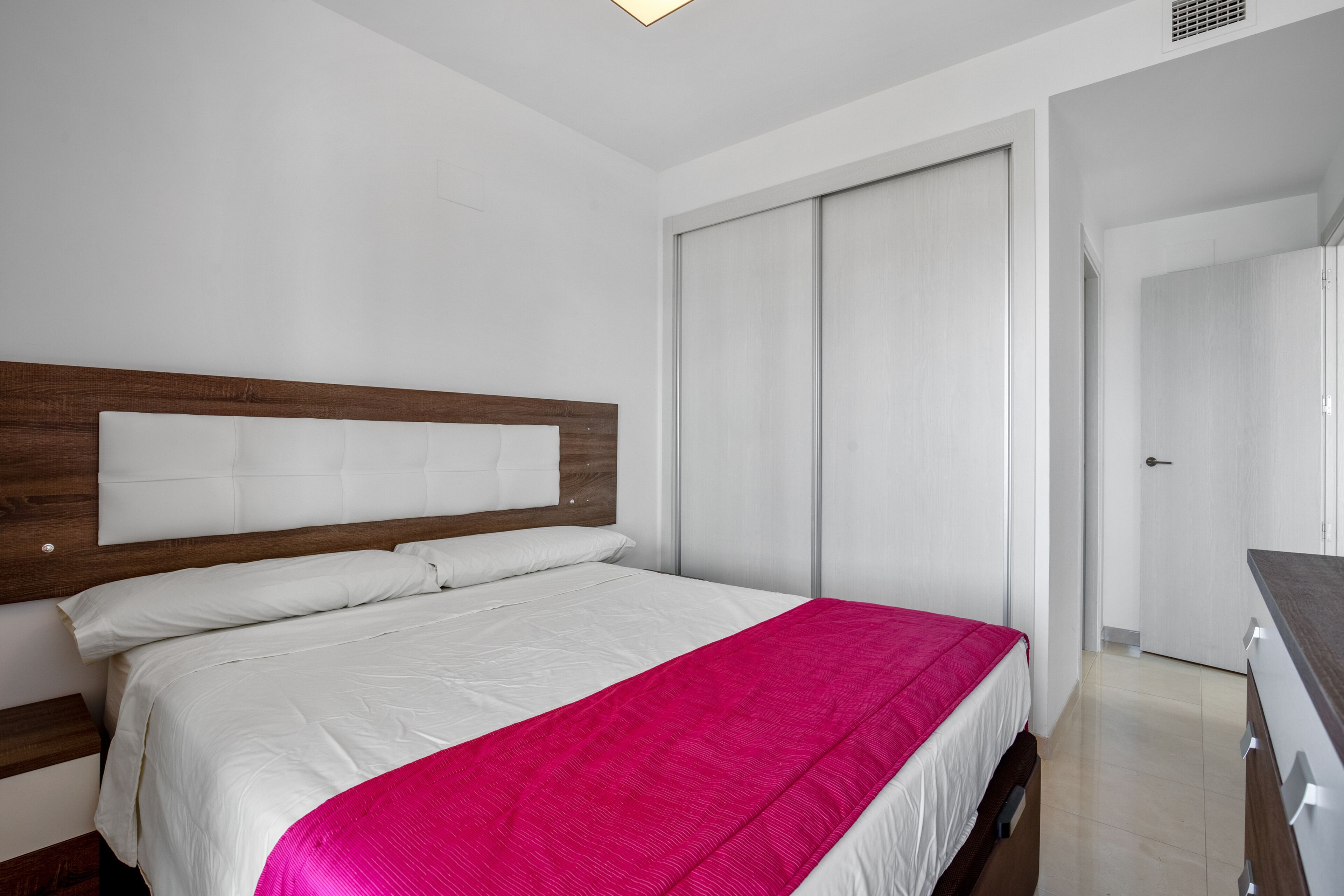 A modern bedroom with a wooden headboard, a white bedspread, and a bright pink accent.