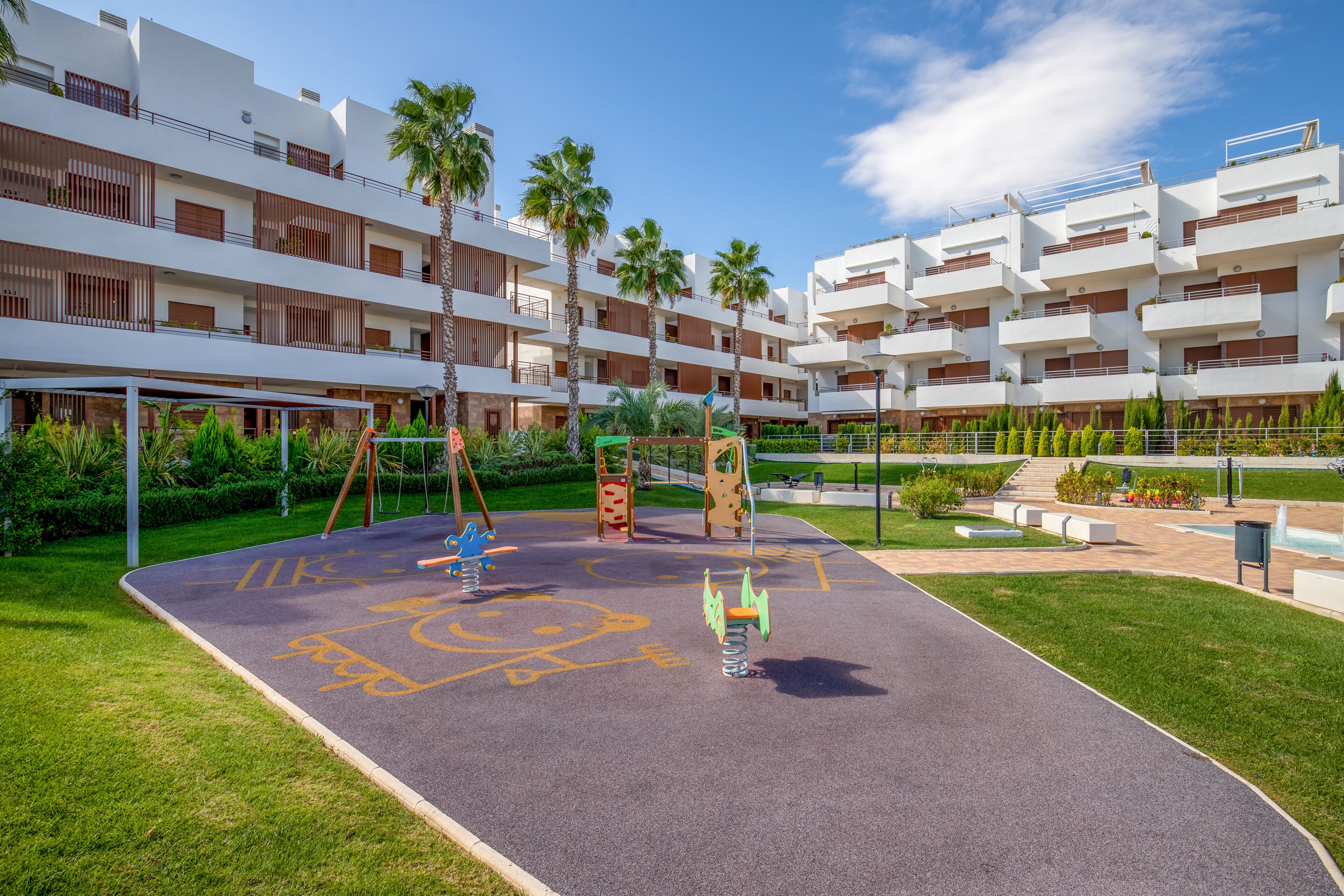 Un parque infantil con columpios y estructuras para escalar, rodeado de un césped cuidado y palmeras, con edificios modernos al fondo.