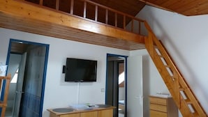 A room with a wooden ceiling, a TV mounted on the wall, a desk with drawers, and a staircase leading to an upper level.