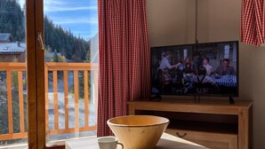 Una habitación con una mesa de madera, una taza blanca, un cuenco de madera, un televisor y un balcón con una cortina a cuadros rojos y blancos.