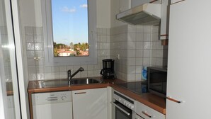 A compact kitchen with white cabinets, a built-in oven, a coffee maker, and a window with a view of houses. - 54m² apartment rental with view of boats, shaded and sunny St Cyprien (Pyrénées-Orientales)