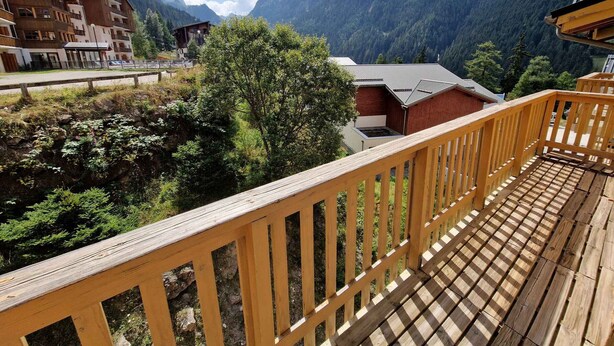 Une terrasse en bois offrant une vue sur un paysage montagneux, un bâtiment doté d’un toit en pente et un arbre au premier plan.