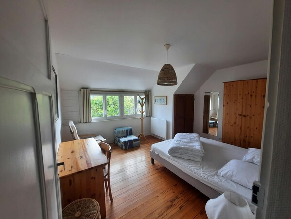 A bedroom with a wooden floor, a bed, a desk with a chair, a window, and a hanging light fixture.