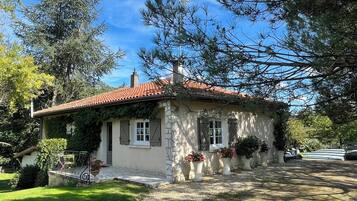 Una pequeña casa de piedra con techo de tejas rojas, rodeada de follaje y árboles.