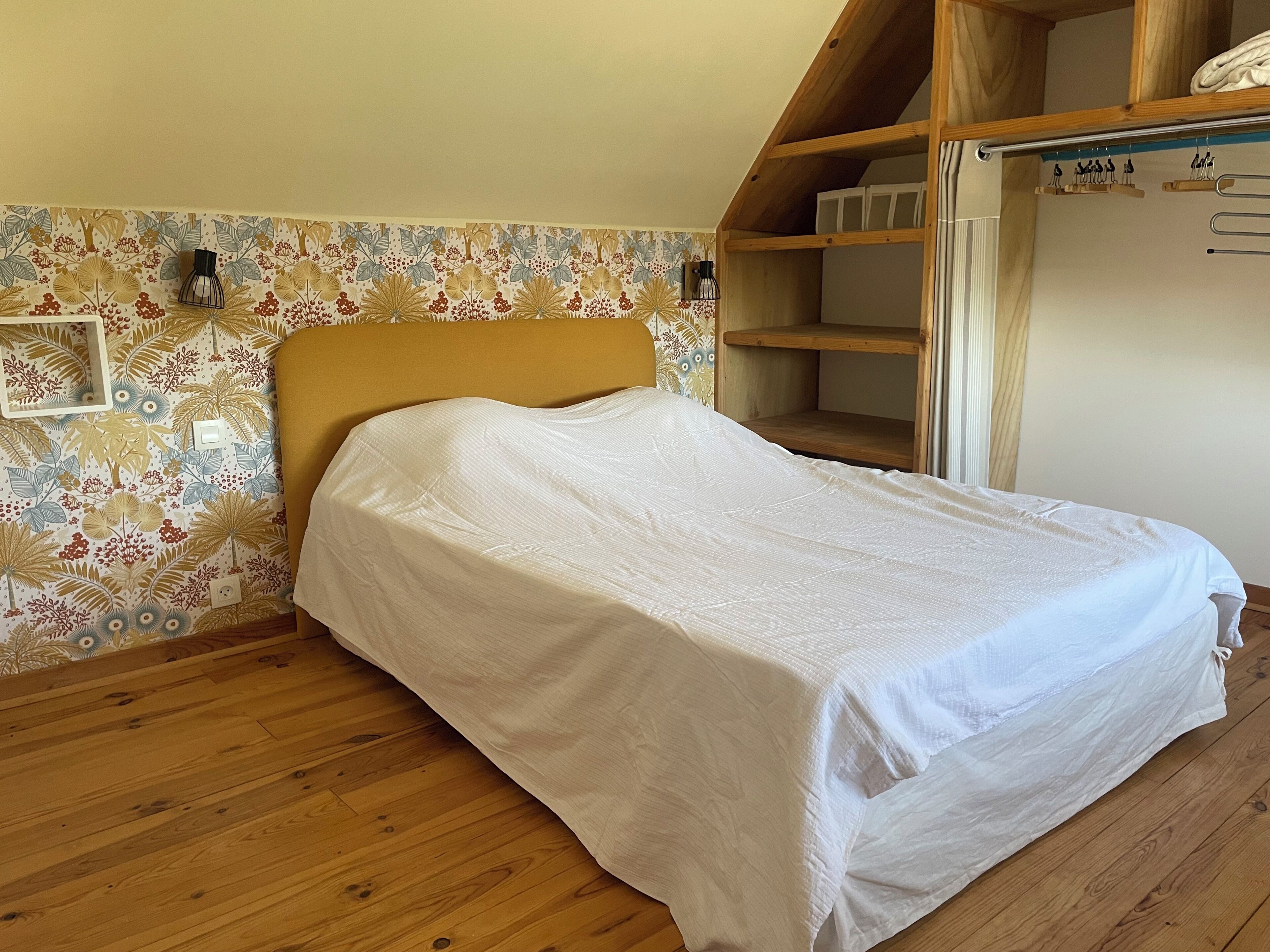 A bedroom with a wooden headboard, a bed with a white cover, a wooden shelf unit, and a wall with a floral pattern.