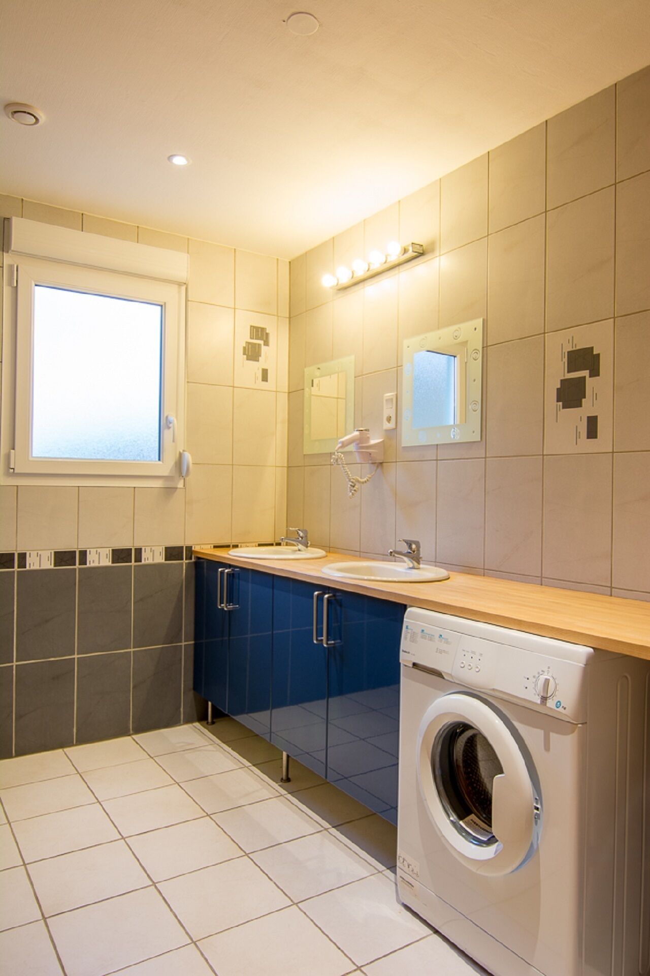 Een kleine badkamer met een ingebouwde wasmachine, een raam met zonwering en een spiegel boven de wastafel.