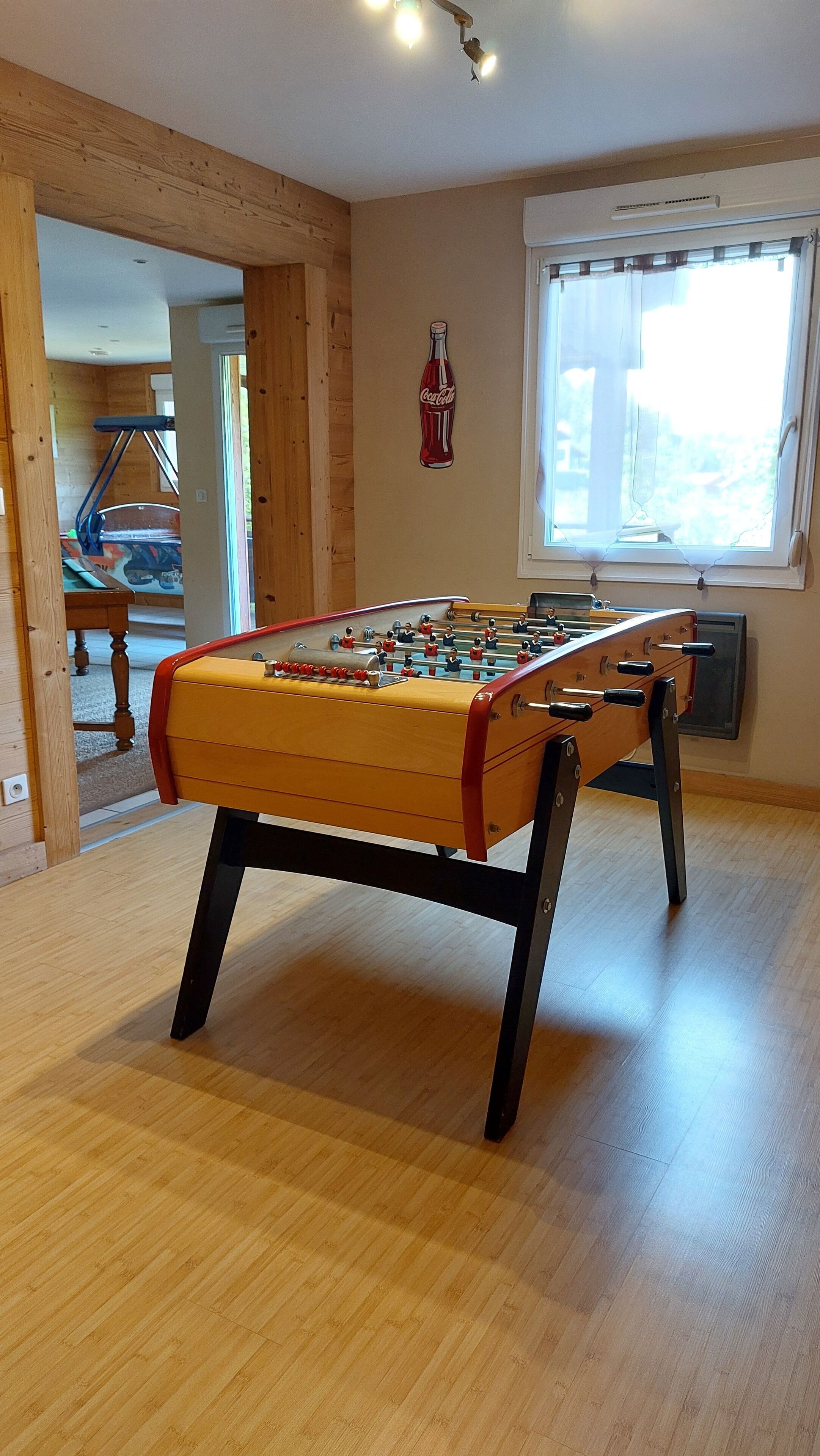 A foosball table in a room with wooden flooring, a window with curtains, and a Coca-Cola bottle decoration on the wall.
