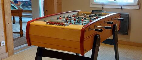 A foosball table in a room with wooden flooring, a window with curtains, and a Coca-Cola bottle decoration on the wall.
