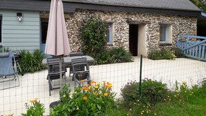 Een stenen huis met een grijs dak, een terras met tuinmeubilair en een hek van gaas.