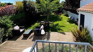 Una terraza de madera con mobiliario exterior, un jardín con césped y un edificio blanco con tejado de tejas.