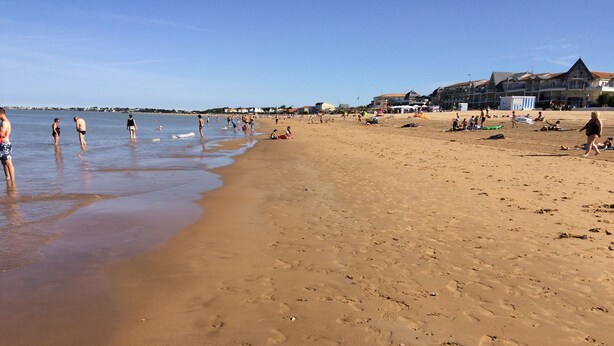 Una playa con huellas en la arena, cielo despejado y edificios al fondo.
